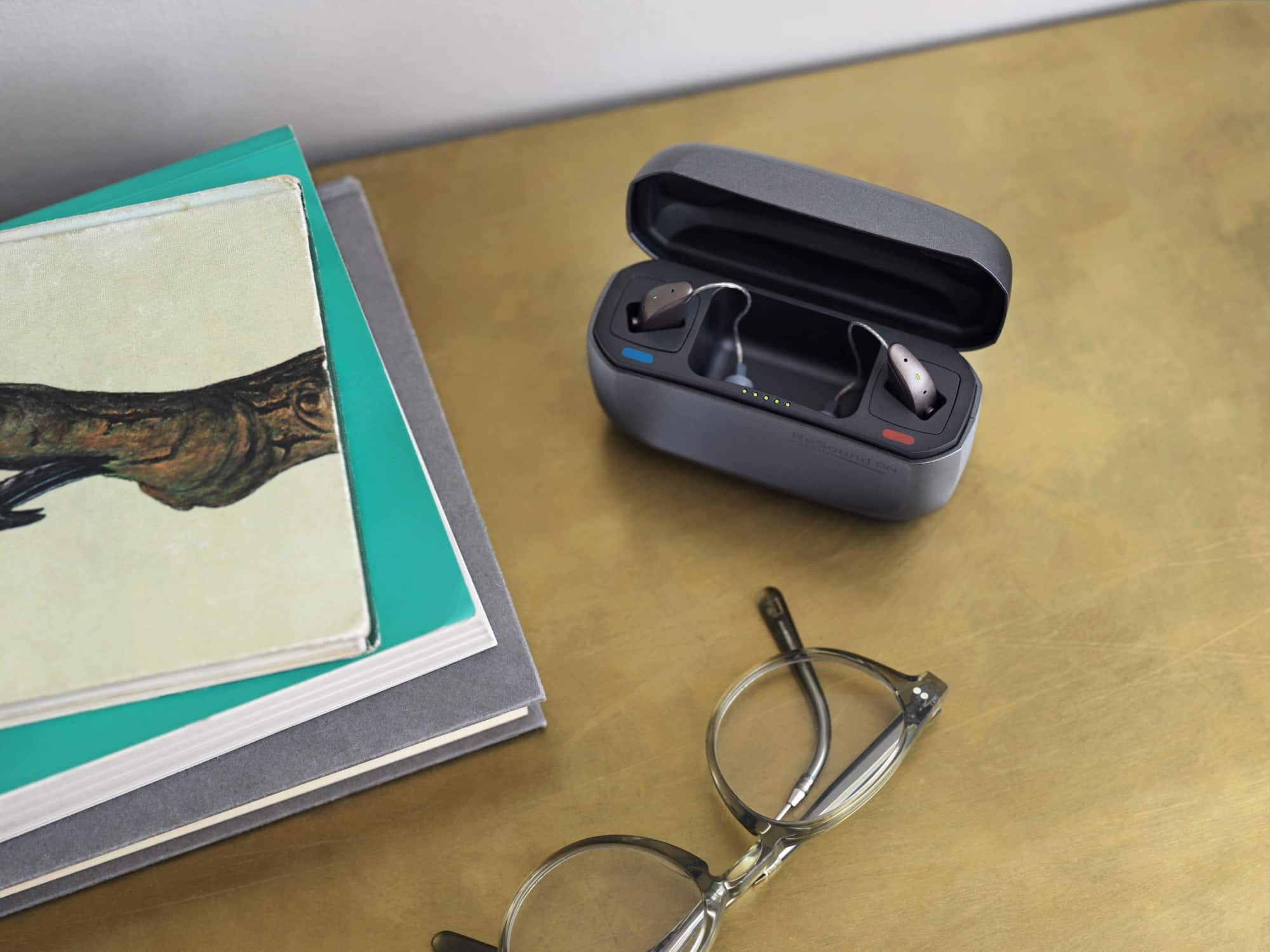 a stack of books, a pair of glasses, and resound hearing aids in a charger